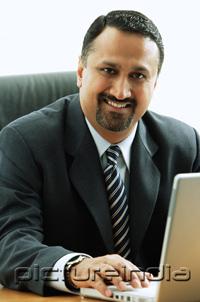 PictureIndia - Businessman at desk, using laptop, looking at camera
