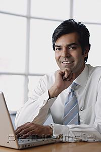 PictureIndia - Businessman sitting in front of laptop, hand on chin, looking at camera