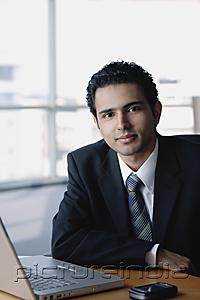 PictureIndia - Young businessman next to laptop, looking at camera