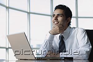 PictureIndia - Young businessman sitting in front of laptop, smiling, looking away